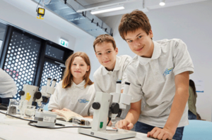 Ein Mädchen und zwei Jungen stehen und sitzen vor Mikroskopen im Schülerforschungszentrum Hamburg.