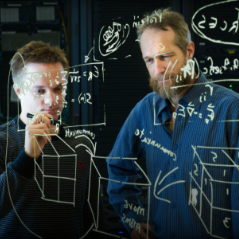 Zwei Männer schreiben mathematische Formel an eine durchsichtige Wand. 