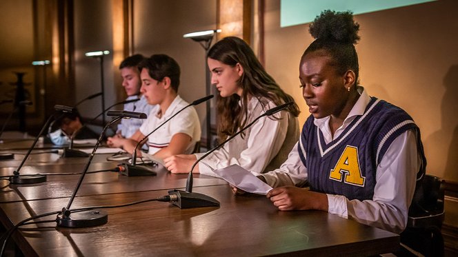 Zu sehen sind vier Schüler:innen, die auf der Bühne an einem Tisch sitzen und vorlesen. 