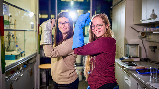 Zwei Personen in einem Labor halten sich große Kissen vor den Gesichtern; im Hintergrund stehen Labortische und Geräte.
