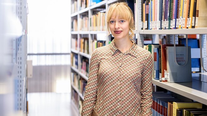 Eine junge Frau steht in einer Bibliothek und lächelt in die Kamera. Es ist eine Alumna aus dem Programm: Janina Steinert.