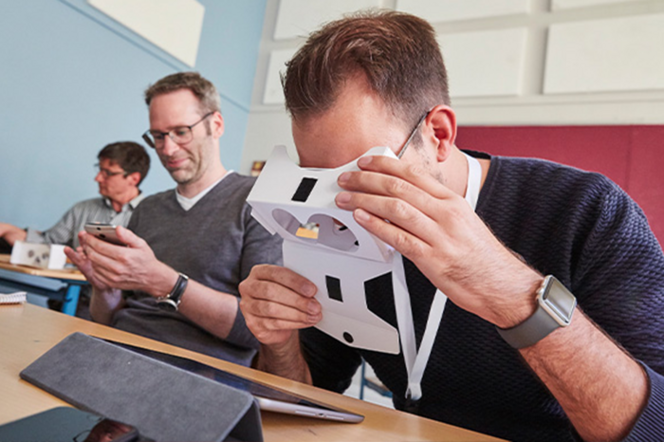 Ein Lehrer schaut durch eine VR-Brille auf ein Tablet. Dahinter schaut ein weiterer Lehrer auf sein Smartphone.