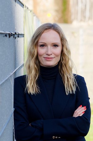 Die Stipendiatin Johanne Düsterbeck, wacher, freundlicher Blick in die Kamera, lehnt an einer Wand und schaut in die Kamera. Sie trägt schwarze Kleidung, hat blonde lange Haare und lackierte Fingernäge.