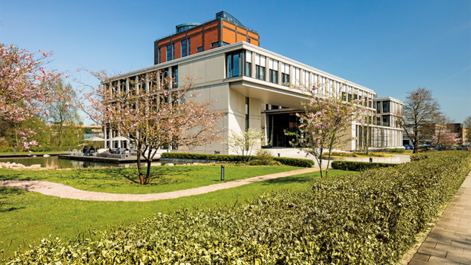 Stiftungsgebäude in Hamburg-Langenhorn. Blick auf das Gebäude und die grüne Hecke drum herum. Es ist eine moderne Architektur, ebenfalls ist der alte Kaffeeturm zu sehen, um den sich das neue Gebäude herumzieht.