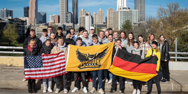 Zu sehen ist eine Gruppe junger Menschen, die vor einer Skyline einer großen Stadt in den USA stehen. Sie halten die deutsche und amerikanische Flagge hoch - in der Mitte die Flagge, des Colleges.