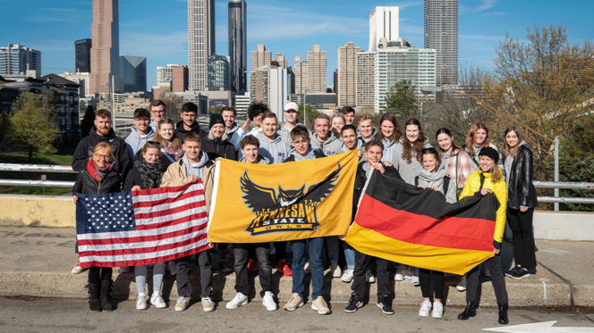 Zu sehen ist eine Gruppe junger Menschen, die vor einer Skyline einer großen Stadt in den USA stehen. Sie halten die deutsche und amerikanische Flagge hoch - in der Mitte die Flagge, des Colleges.