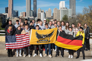 Zu sehen ist eine Gruppe junger Menschen, die vor einer Skyline einer großen Stadt in den USA stehen. Sie halten die deutsche und amerikanische Flagge hoch - in der Mitte die Flagge, des Colleges.