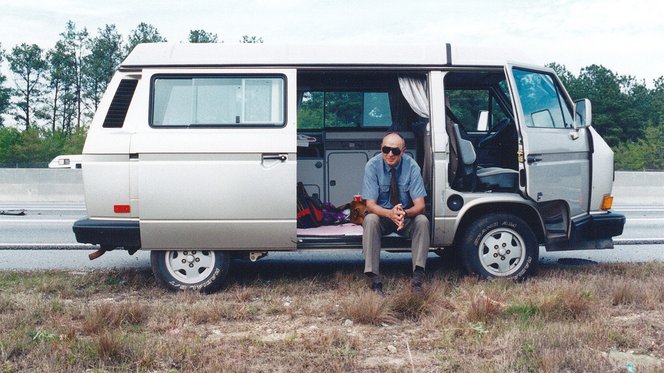 Zu sehen ist ein alter VW-T3-Campingbus in einer grau-metallic-Farbe. Joachim Herz sitzt im Zwischenraum und trägt eine Sonnenbrille. Er schaut direkt in die Kamera.