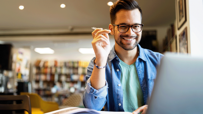 Ein Mann sitzt in einer Bibliothek vor einem Laptop, hält einen Stift in der Hand und lächelt in die Kamera. 