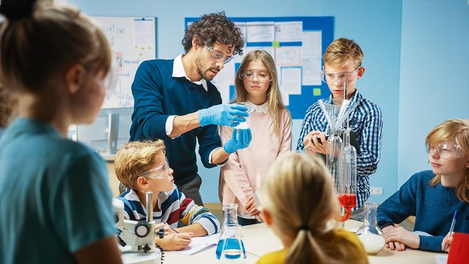 Ein Lehrer hält einen Behälter eines Experiments hoch, um ihn seinen Schüler:innen zu zeigen. Alle tragen Schutzbrillen.