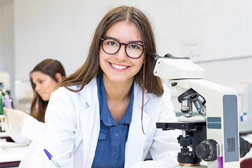 Eine Add-on Fellow im Laborkittel blickt über ihr Mikroskop hinweg in die Kamera