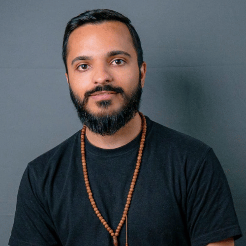 Portrait Ali Alam, he wears a dark T‑shirt with a beaded necklace against a gray background