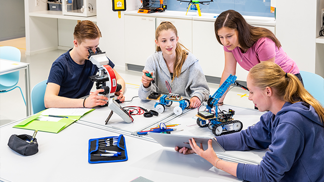 Drei Schülerinnen und ein Schüler sitzen und stehen um einen Tisch, auf dem selbstgebaute Fahrzeuge stehen. Die Schülerinnen schauen auf ein Tablet, der Schüler in ein Mikroskop. 