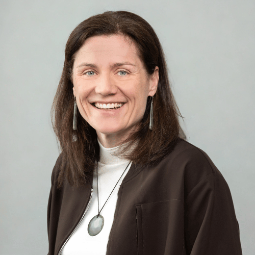 Portrait of a Tina Menker, wearing a light turtleneck, dark jacket and pendant against a light background
